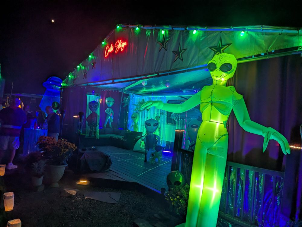 A deck lit with green and blue lights, decorated with various inflatable big head aliens. A large translucent green alien stands to the right of the deck