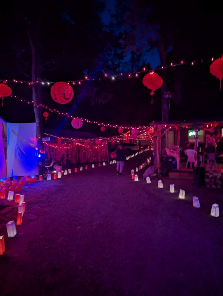 A pathway lined with red and white luminaries, with an overhead crisscross of red and white lights. Round red paper lanterns of various designs hang from the lights