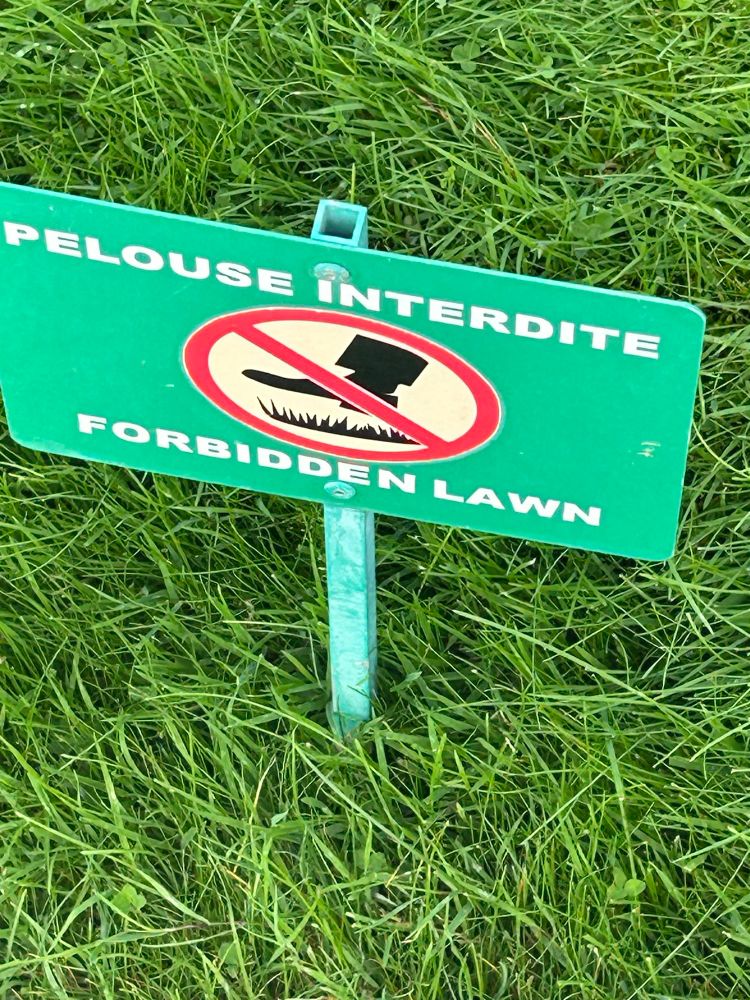 A green sign indicating you should not walk on the grass, in both French and English. The French is Pelouse Interdite, the English is Forbidden Lawn. Beneath the sign is a section of shortish cut grass.