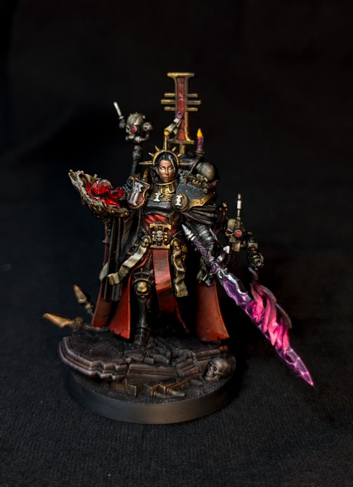Miniature of a woman inquisitor from 40k, armored in black and gold with robes of red and black 
She holds a book of heretical texts with a shadowy hand emerging from it and a daemonic longsword with a purple blade, emitting magenta flames 