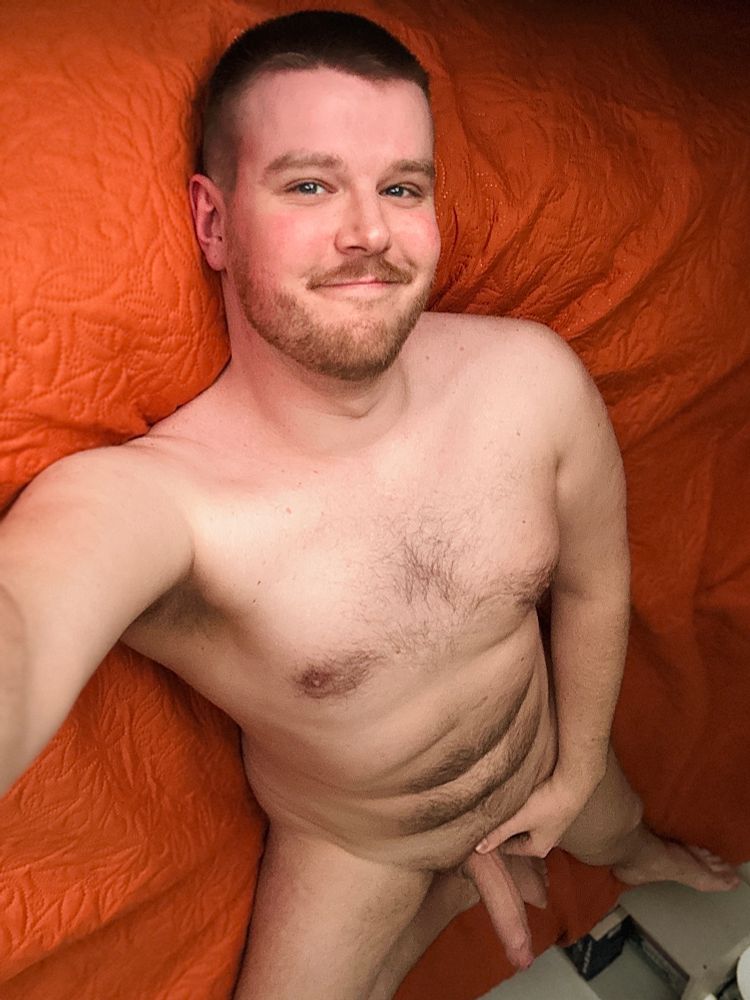 Naked man on orange bedsheet taking selfie while laying down