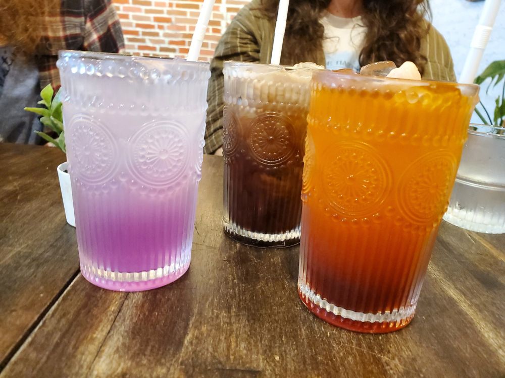 3 drinks in clear glasses lined up on a table; one drink is purple, another brown, the third orange.