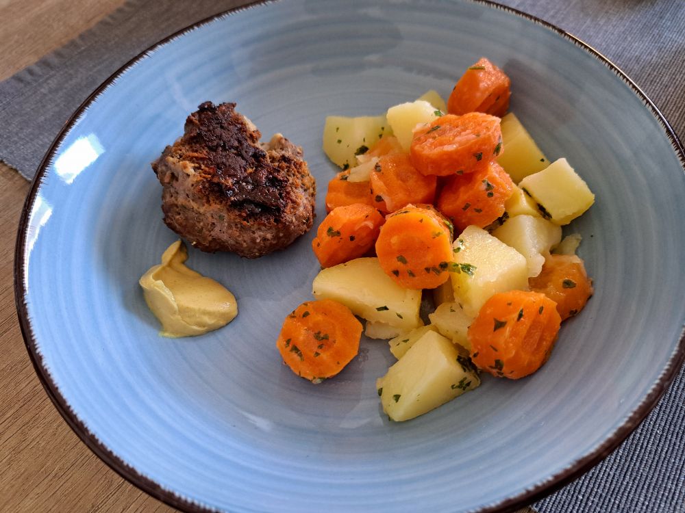 Eine Frikadelle mit nem Klecks Senf auf einem Teller mit Kartoffeln und Möhren. 