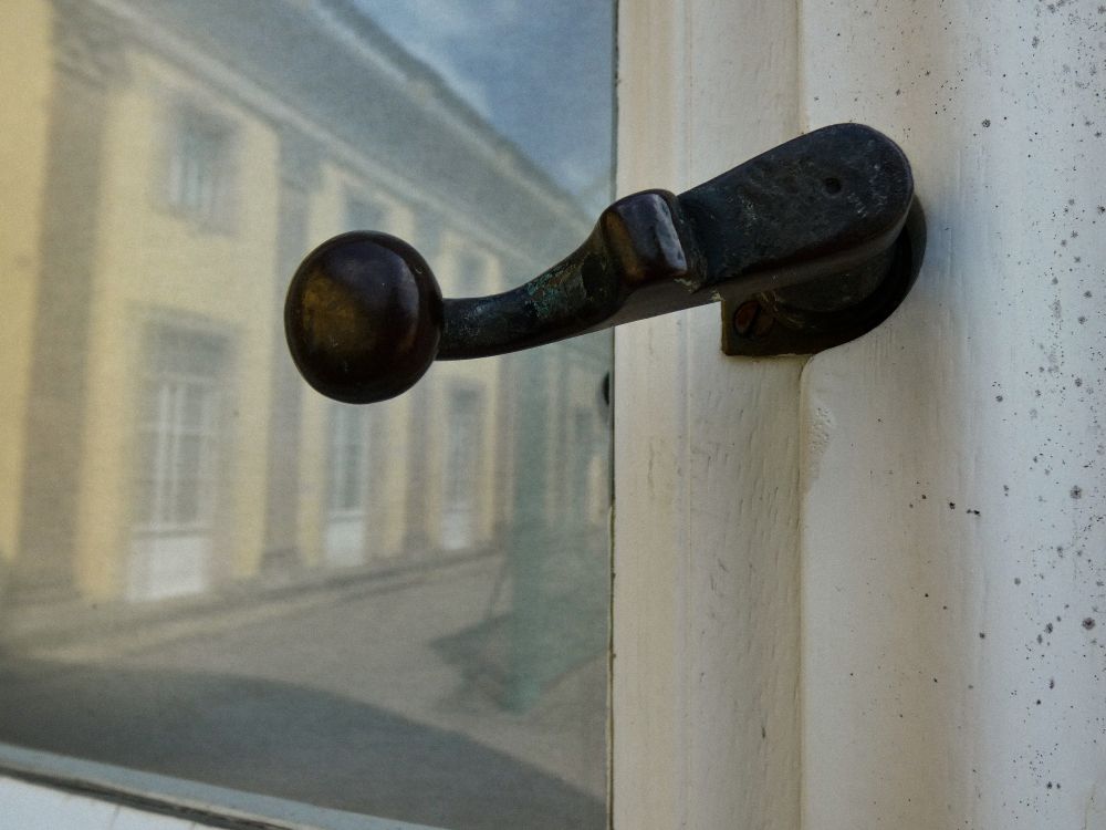 Ein schwarzer Türgriff an einer weißen Tür. In der Glasscheibe spiegelt sich ein Teil des Schlosses Sanssouci 