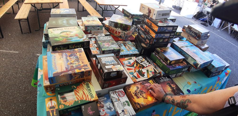 La table avec les jeux un peu plus exigeants tels que Heat, Akropolis, Demain je t'ai tué...etc. sur la table une bonne cinquantaine de jeux empilés les uns sur les autres. On aperçoit le bras tatoué de mon amie Claire en train de regarder les jeux.