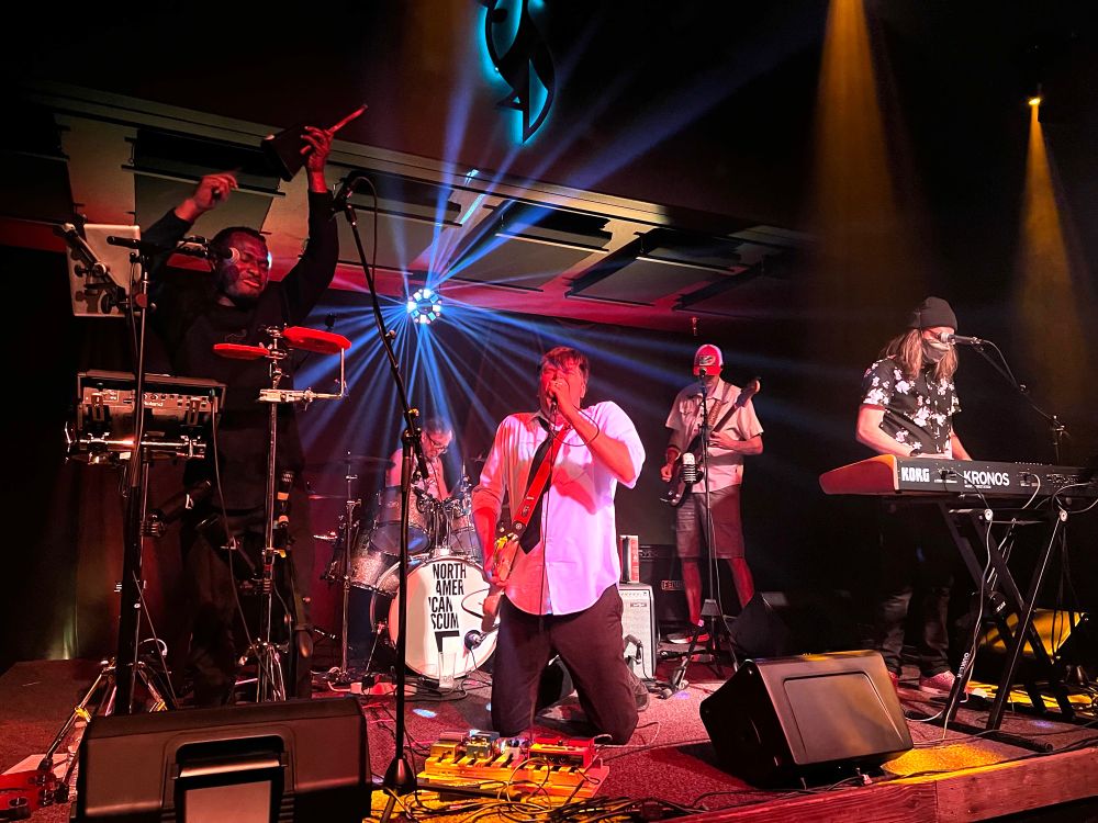 singer wearing a white shirt on his knees with other musicians next to him on stage (percussion, drums, bass, keyboards). 