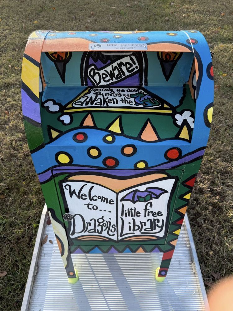 Colorful mailbox painted with a dragon theme and the words 'Welcome to Dragon's Little Free Library' on the front panel.
