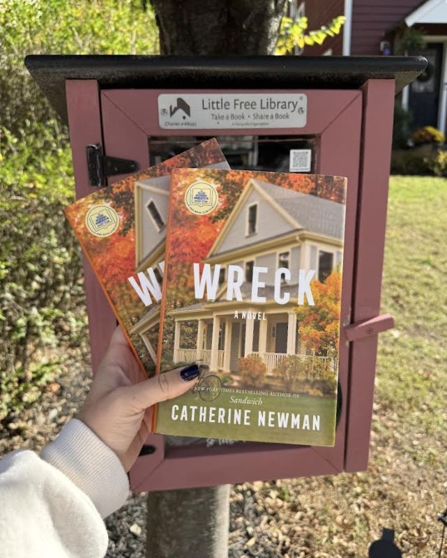 Hand holding two copies of the novel 'Wreck' by Catherine Newman in front of a Little Free Library outdoors on a sunny day.
