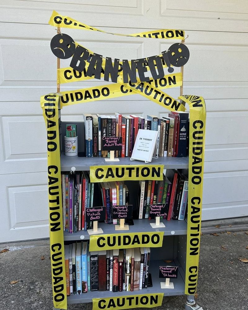 Bookshelves wrapped in yellow caution tape with signs labeling sections as challenged or banned adult, children's, graphic novels, picture, and YA books.