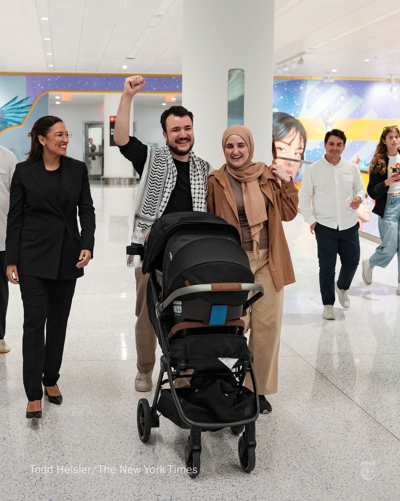 Mahmoud Khalil, wearing a keffiyeh, pushing a stroller and walking between his wife, Dr. Noor Abdalla, their Alexandria Ocasio-Cortez. His hand is raised in a fist and all three people are smiling. Photo by Todd Heisler/The New York Times