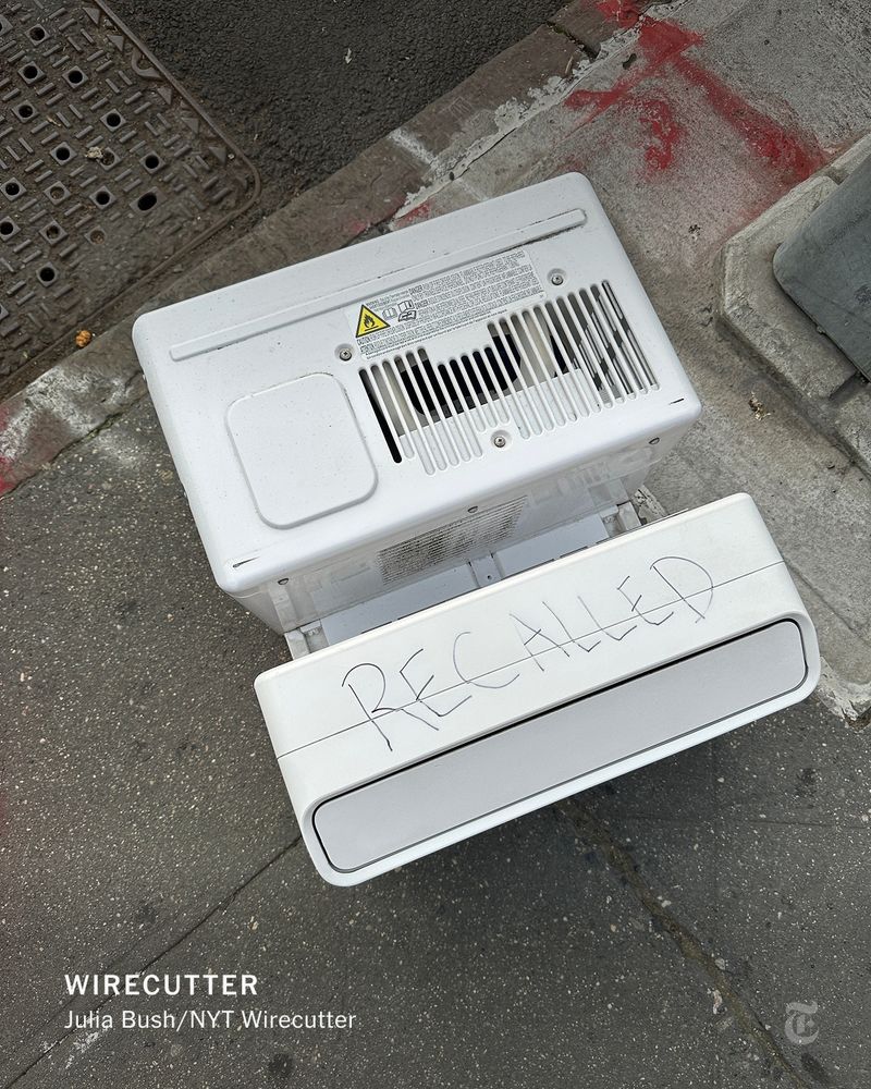 A white, disassembled air conditioner with the word "RECALLED" written on it in black marker, resting on a gray sidewalk next to a metal grate.