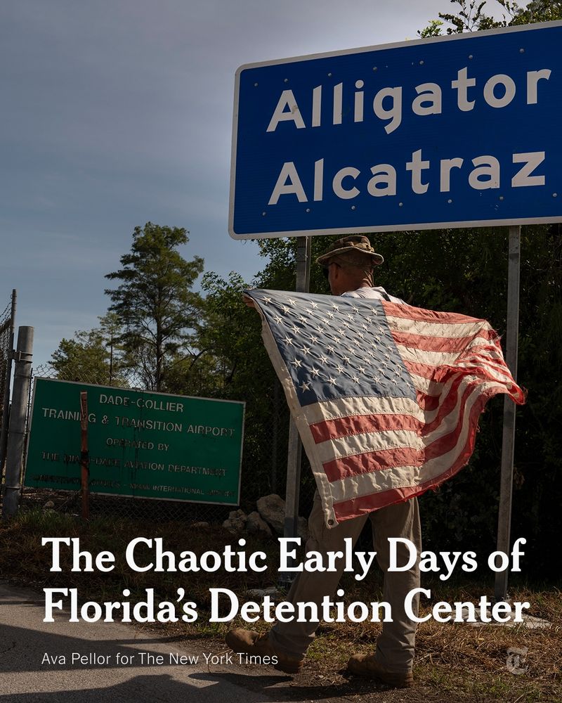 A blue sign with white writing says “Alligator Alcatraz.” A man stands in front of the sign facing away from the camera, holding a faded American flag across his back. A headline reads: “The Chaotic Early Days of Florida's Detention Center.” Photo credit: Ava Pellor for The New York Times.