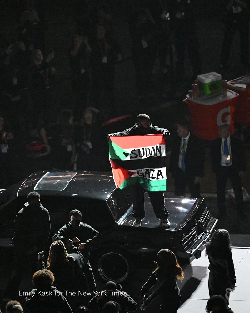 A person stands on a black car holding a green, black, red and white flag with the words “Sudan” and “Gaza” on it. Dozens of people are dancing onstage nearby. Photo credit: Emily Kask for The New York Times.