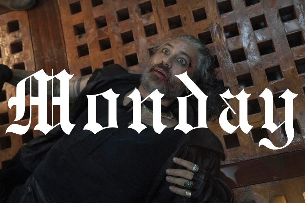A screenshot from Our Flag Means Death season 2, episode 2. It shows Taika Waititi as Ed, laying with his back on the Revenge's deck, wet from the rain. His looks shocked and has various bleeding injuries in his face. Overlapped in the picture is the word "monday" in white gothic letters