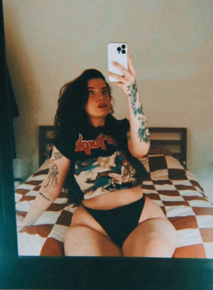 a mirror selfie of anni - a white woman with long dark hair wearing a “deep purple” tshirt. you can see the tattoos on her arms and she’s sitting on her bed 