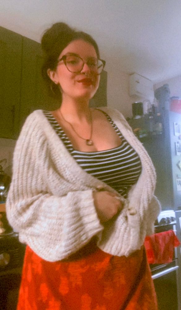 a selfie of anni, a white woman with dark hair in a bun on top of her head and big glasses. she is wearing a big cream coloured cardigan, an orangey red floral skirt and a stripe top. 