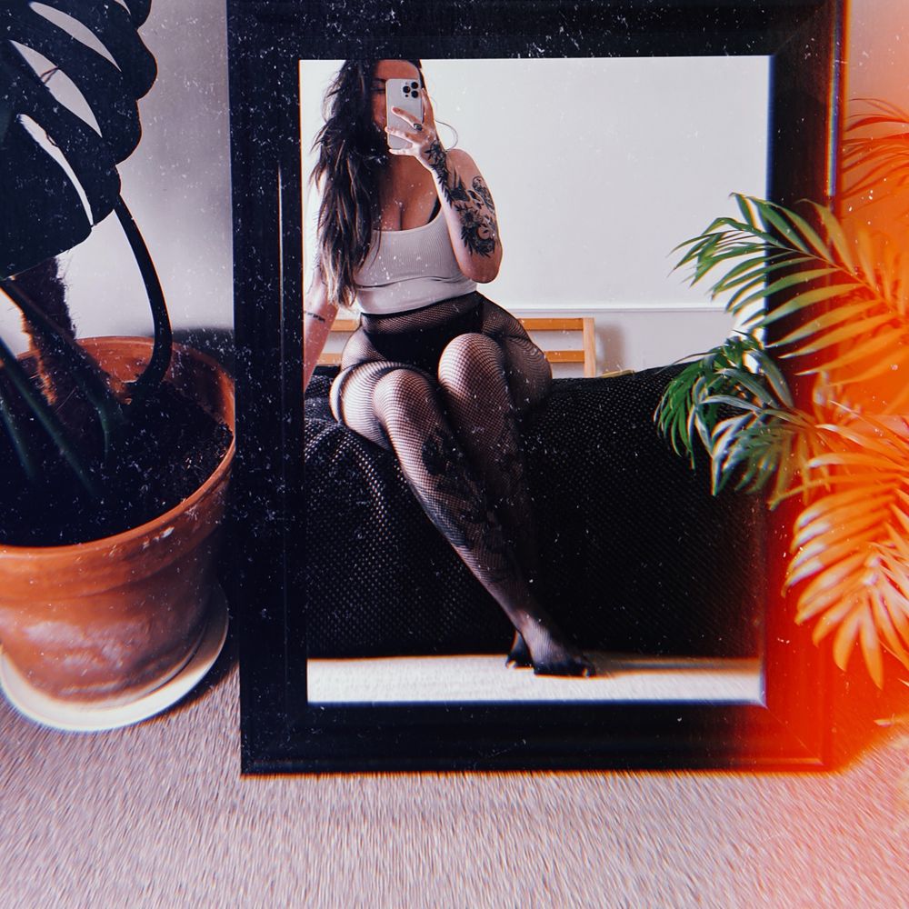 anni taking a mirror selfie with plants at either side of the mirror, she’s wearing a white crop top and fishnets, her long dark hair is down and her phone is covering her face