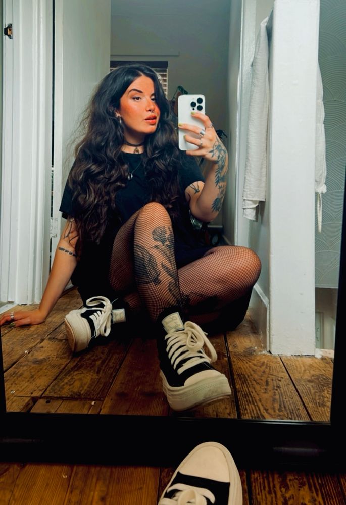 a mirror selfie of anni, a white woman sitting on a hardwood floor. she is wearing a big black tshirt, fishnet tights and black converse. she has tattoos on her arms and legs and she is also wearing a choker around her neck. she has long thick dark wavy hair over her shoulder. 
