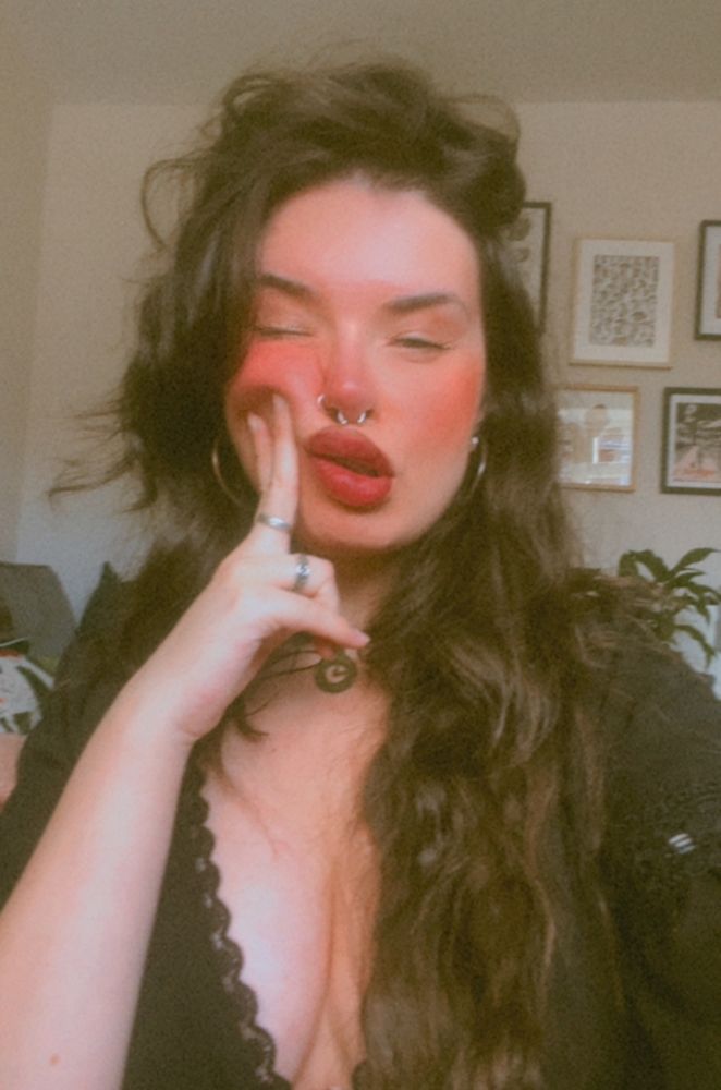 a selfie of anni wearing a black dress, her long dark wavy hair is pushed over the side and hanging over her chest. she is squinting and pushing two fingers against her rosy cheek. 