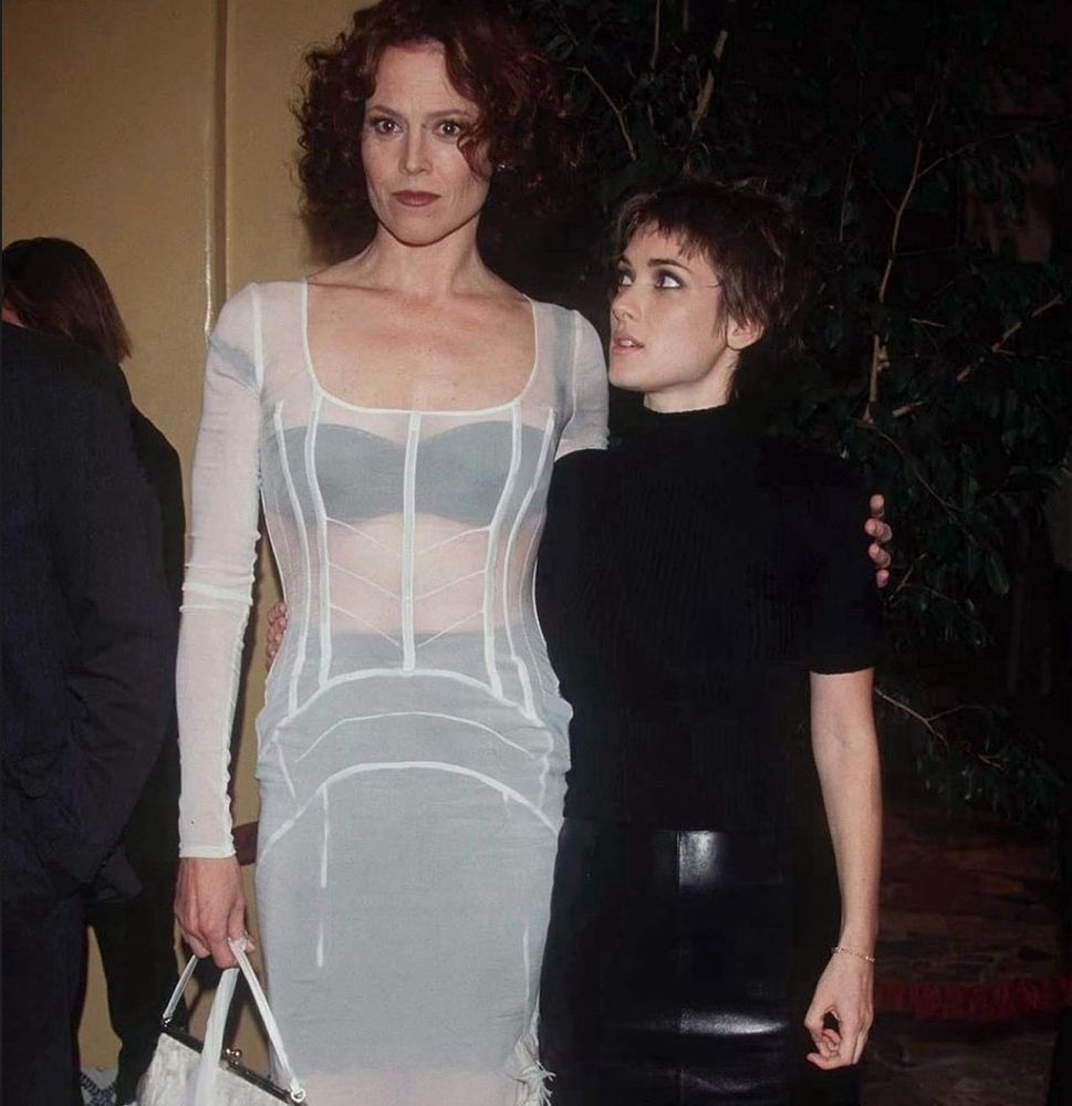 sigourney weaver and winona ryder - a tall white woman on the left with short dark hair in a white dress with her arm around a shorter white woman on the right with short dark hair, the woman on the right is gazing up at her while the woman on the left looks at cameras pointed at her