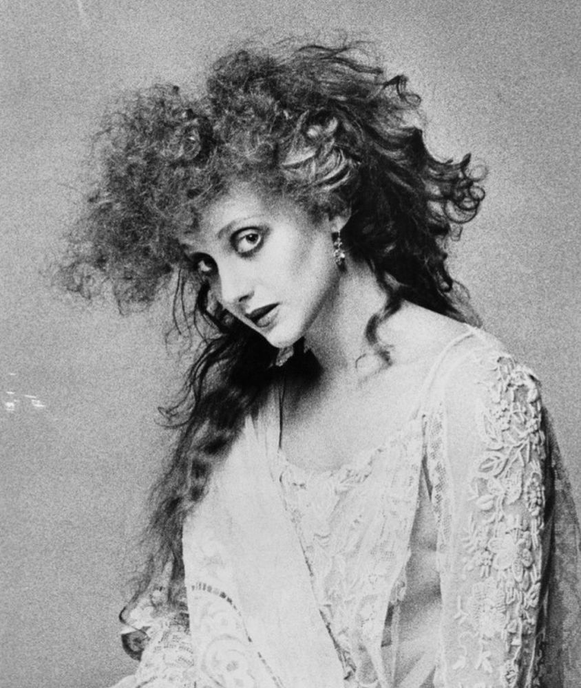 a black and white photo of carol kane from 1976, she is wearing a white lace robe and has fluffy big hair. 