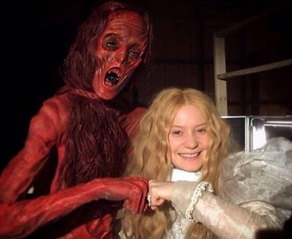 a white woman with blonde wavy hair wearing a puffy dress is fist bumping a tall red ghostly woman creature 
