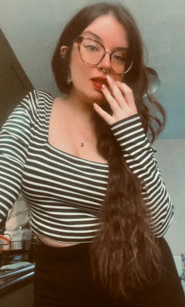 a selfie of anni, a white woman with long dark wavy hair hanging over her shoulder and her chest. her hand is against her mouth and cheek. she is wearing large turtle shell glasses, a black and white striped crop top and a black skirt. 