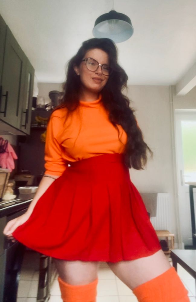 anni (a white woman with thing long dark wavy hair and glasses) is dressed as velma with an orange turtleneck sweater, a red skirt and orange thigh high socks. she is standing in her kitchen smiling 