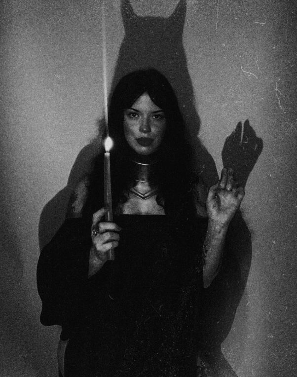 a black and white portrait of anni, a white woman with long dark hair over her shoulders. she is wearing a black, off the shoulder dress with poofy sleeves a silver necklace. in one hand she’s holding a lit candle and the other hand is resting palm facing upwards. her shadow behind her has devil horns. 