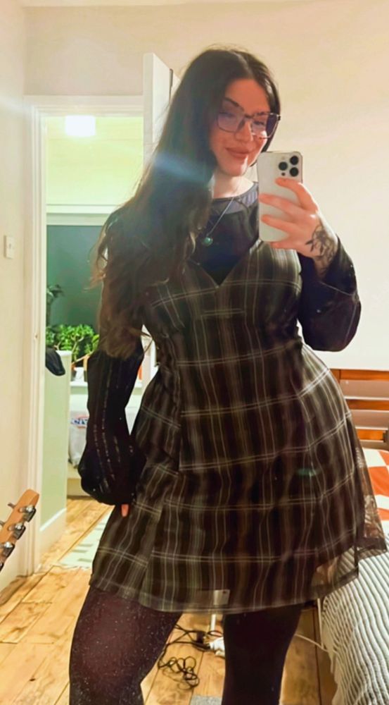 a mirror selfie of anni wearing a green checked dress and a long floaty sleeved black top underneath, she is also wearing large framed glasses and her long dark hair is over her shoulder 