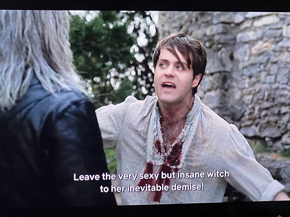 a man in a white blouse covered in blood exclaiming to a man dressed in black leather with white hair “leave the very sexy but insane witch to her inevitable demise!” 