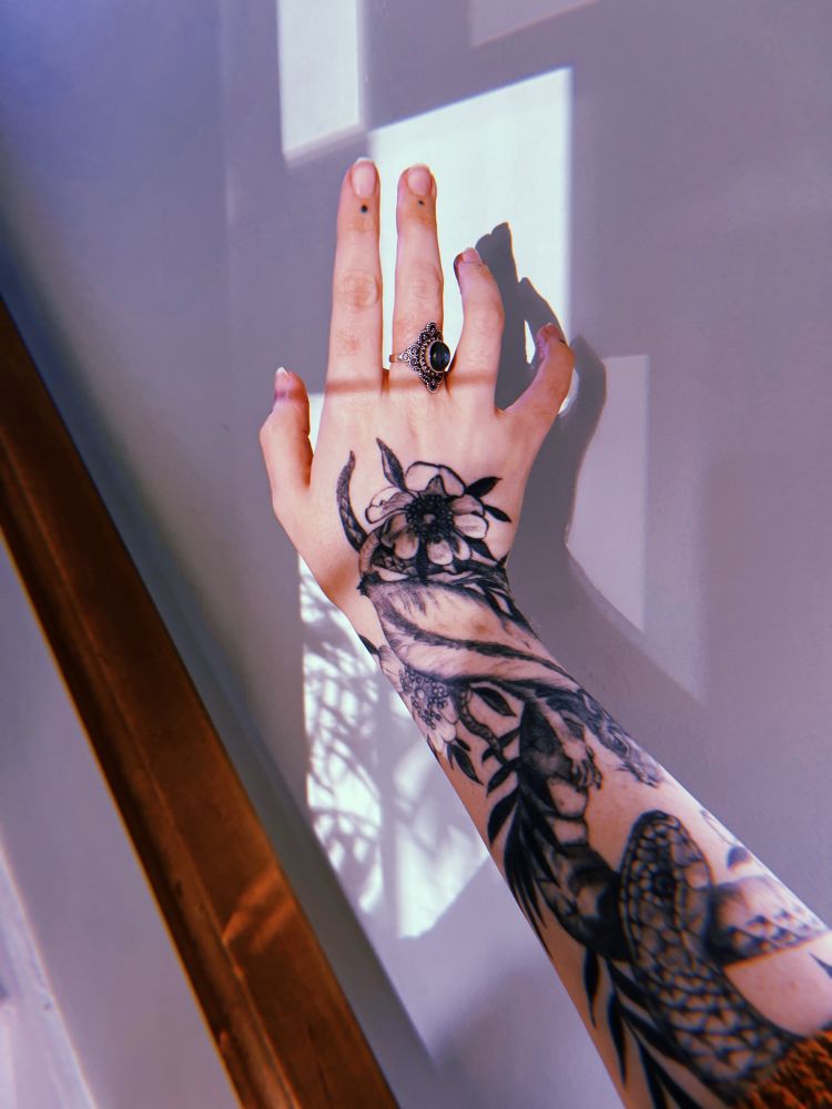 a tattooed arm and hand with an ornate ring on the middle finger, sunlight making shadows on the wall in the background. 