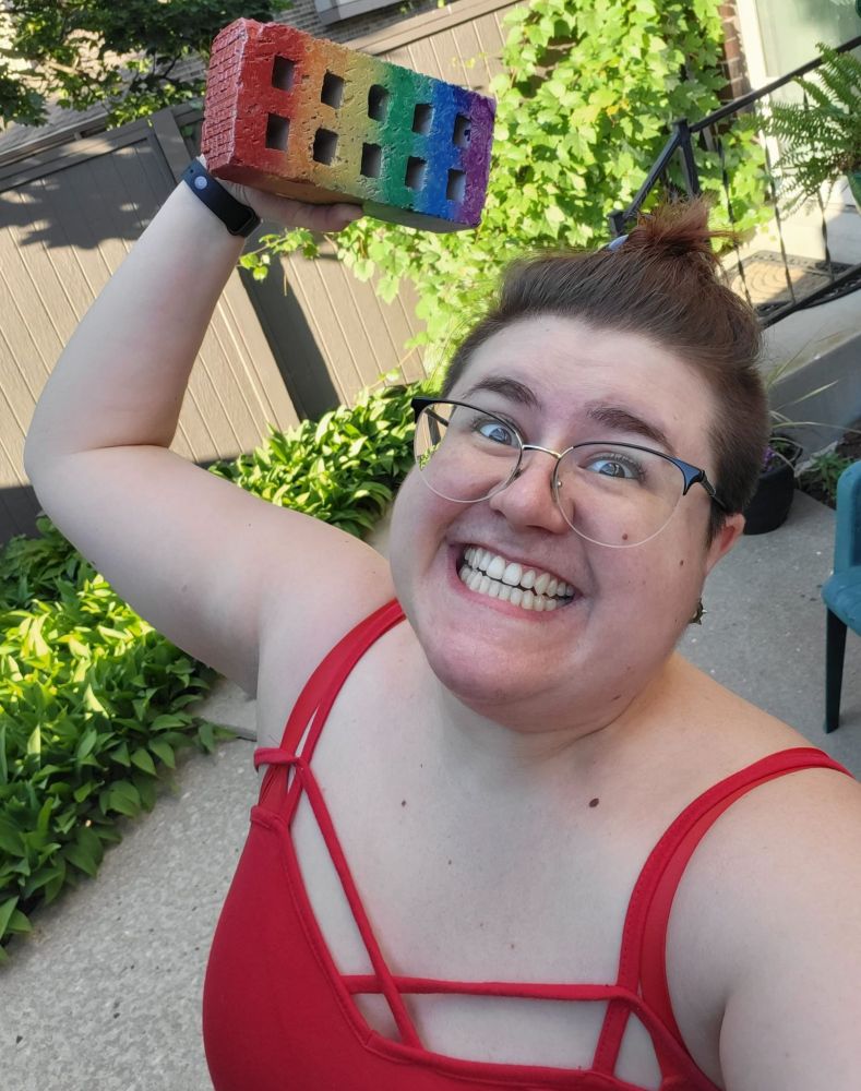 Selfie of me in a red tank top holding up a brick spray-painted in rainbow colors with an unhinged smile on my face.
