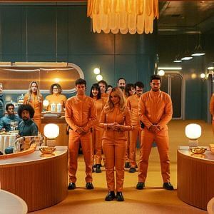 Un puñado de gente vestida con monos naranja en una sala estilo años 50 con paredes turquesa y habitantes del refugio sentados, estos con monos también turquesas. 