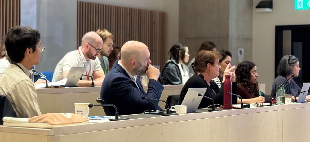 A row of professionals sitting in conference chairs. The individuals all face forward with listening facial expressions.
