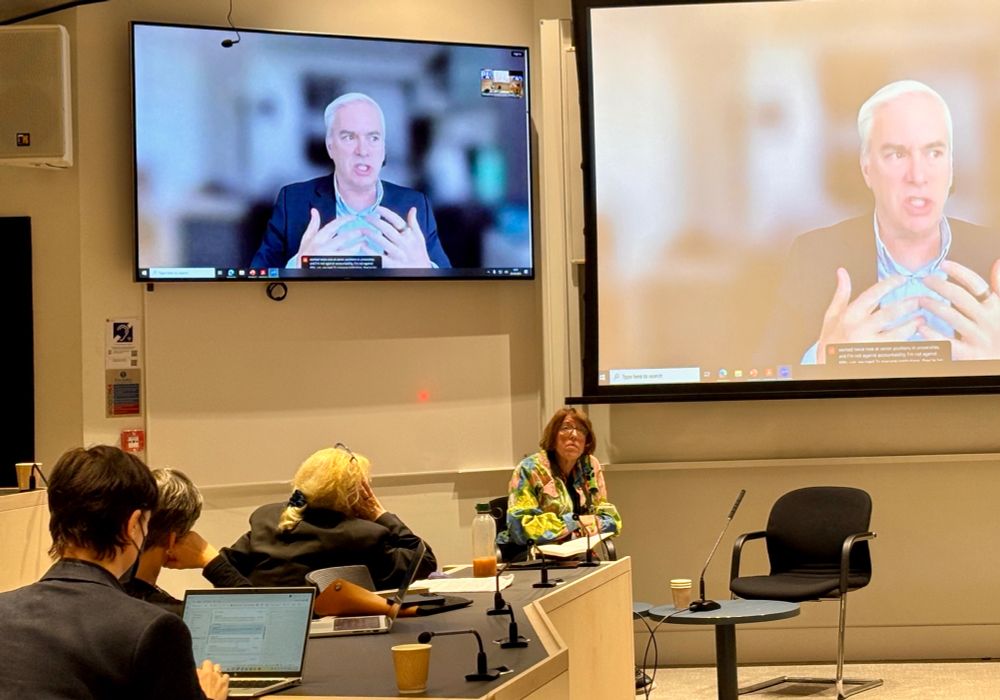 A man speaking appears on two large screens mounted to the wall of a conference room. Audience members sitting in chairs look up at the screen.