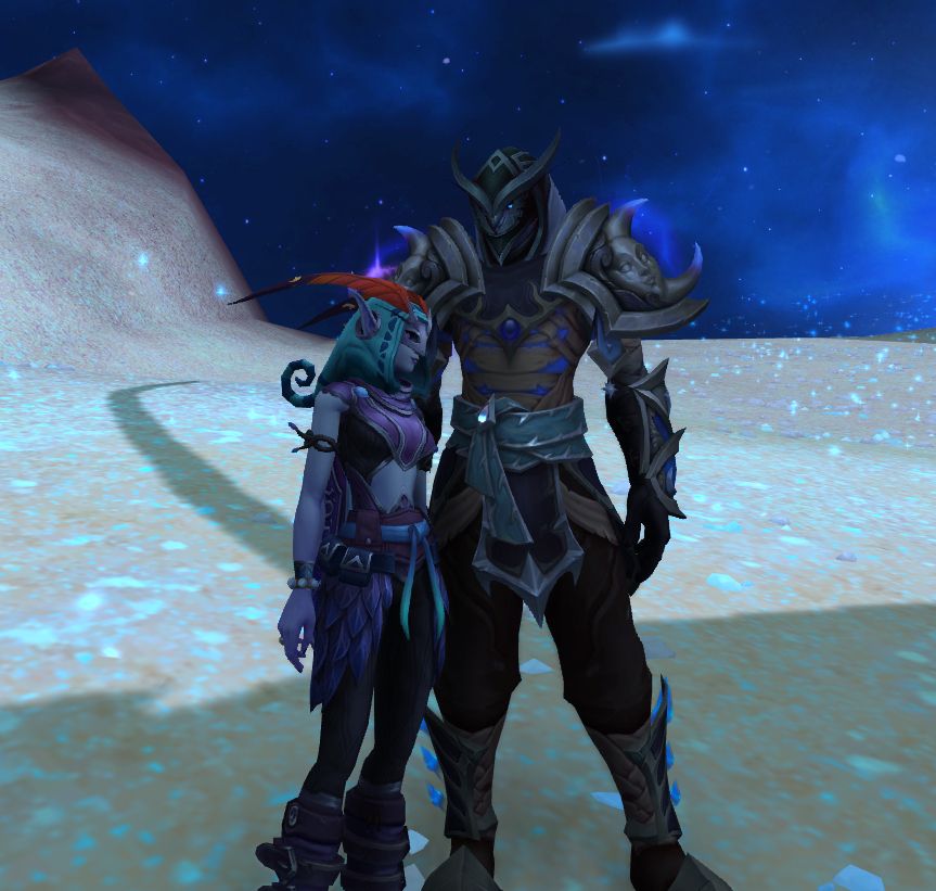Screenshot from World of Warcraft. Two masked figures stand on glittering sand, the woman dressed in mothlike antennae and a wing-shaped cloak, the man with an owl mask. She wears primarily violet and pale blue, her skin ghostly and almost sickly in tone; wearing black and teal-green bound in silver, he shows no visible skin, but the bluish light of energy peeks out from between the wrappings on his torso, revealing that he is likely an Ethereal.