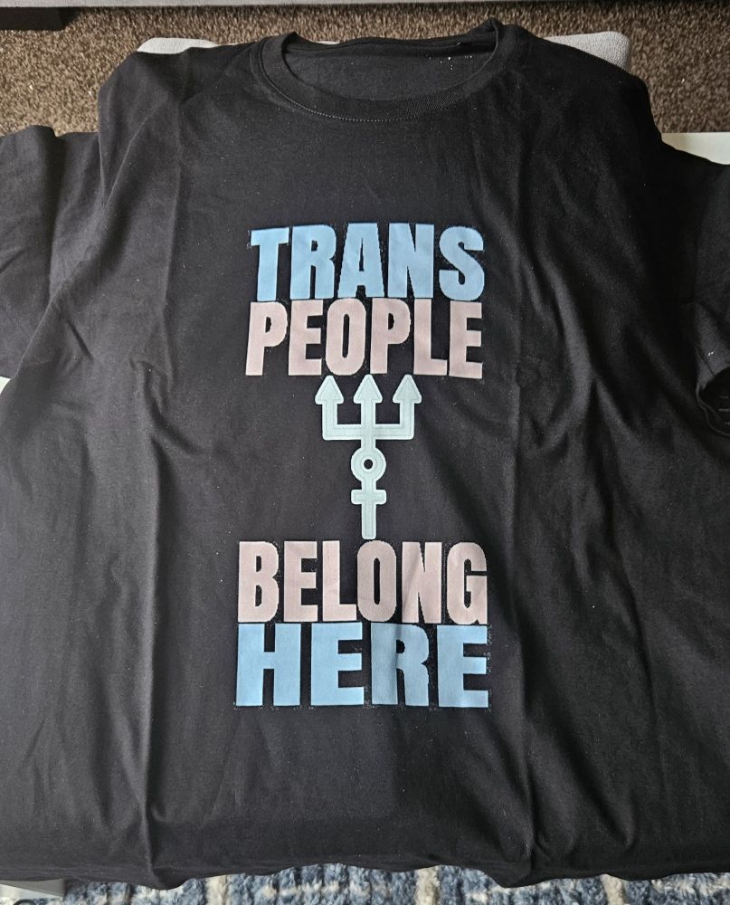 A black t-shirt with the words "trans people belong here" the words are stacked above and below the white Proteus Trident in the colours of the trans pride flag