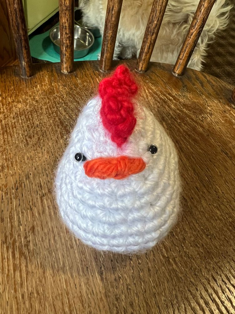 A white crochet chicken with red comb, orange beak, and black eyes on a chair. 