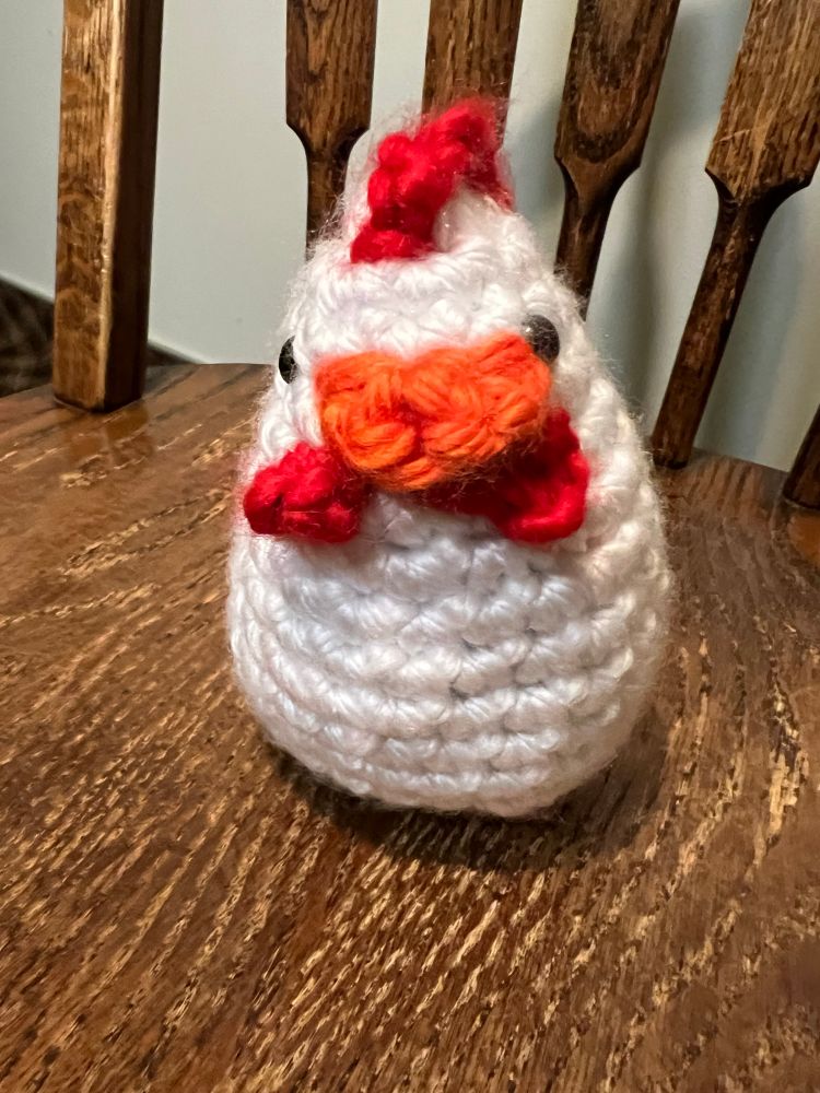 Photo of crochet white chicken with red comb, yellow beak, and red woddles of chicken facing forward.