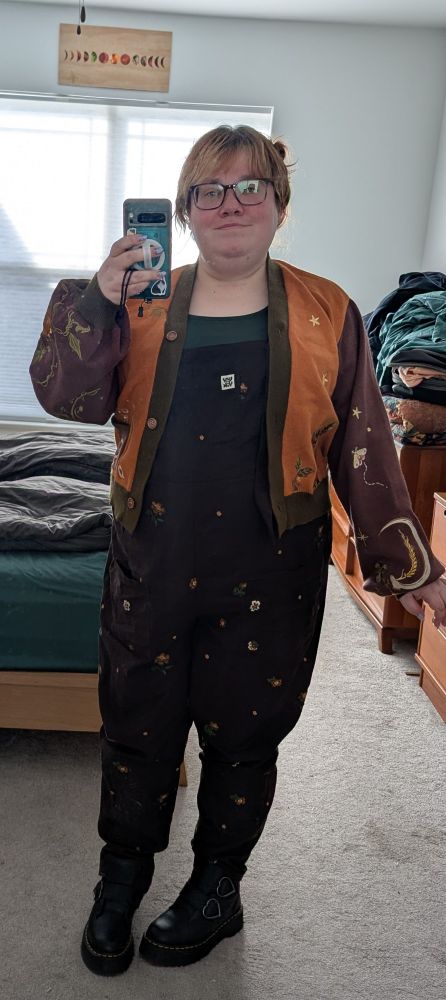 Full length selfie of a nonbinary person taken on their cell phone standing in front of a mirror in their bedroom. They're wearing an embroidered cardigan sweater in orange, green, and brown over brown dungarees embroidered with flowers and black platform boots with heart buckles. Their hair is up in space buns and they're wearing large tortoiseshell glasses.