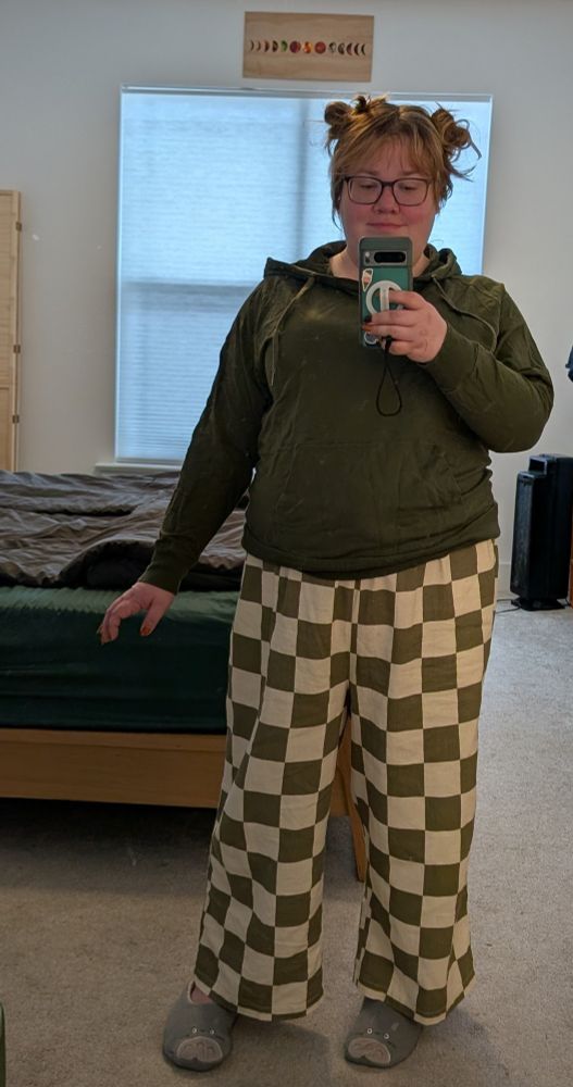 A photo of a nonbinary person taken as a full-length selfie in the mirror. They have red-brown hair tied up in space buns, and bangs, and large tortoiseshell framed glasses. They're wearing an olive green hoodie and wide leg pants with a large olive green and cream checkered pattern and fuzzy gray socks with Totoro on the toes.