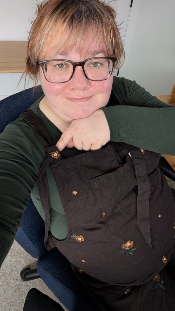 A selfie taken of Wren, a nonbinary person sitting in a desk chair wearing large tortoiseshell framed glasses. They have reddish brown hair with bangs and the rest of the hair is currently styled in space buns. They are wearing a green long sleeve tee and chocolate brown dungarees with flowers embroidered on them. They're pointing at a flower on one of the strap ties, which is positioned perfectly to the front of the tie.