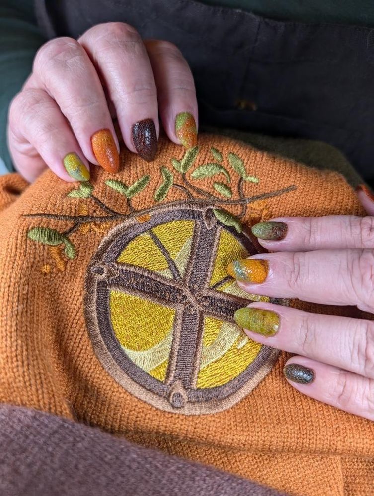 A photo of hands on a part of the sweater, posed to show the nail art. The fingernails are oval and colored in blended orange, light green, and brown, with a scaled pattern on top. The colors of the nails match the sweater and its embroidery, orange, brown, yellow, and green. The embroidery shown in this picture is of a round window divided into fourths with a brown frame and yellow inside. A green leafy branch is embroidered outside the window.