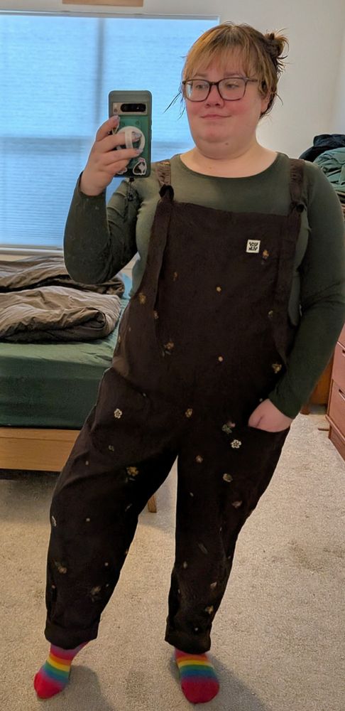 A full-length selfie taken in front of a mirror in a bedroom by a non binary person, Wren. They're wearing a forest green long sleeve T-shirt under chocolate brown dungarees that are embroidered with small flowers in shades of orange and tan. The dungaree straps tie at the chest in place of metal hooks. They're also wearing rainbow socks, and their reddish brown hair is up in two space buns. They're wearing large frame tortoiseshell glasses and standing with one hand in a hip pocket and one hand holding up the phone taking the selfie.