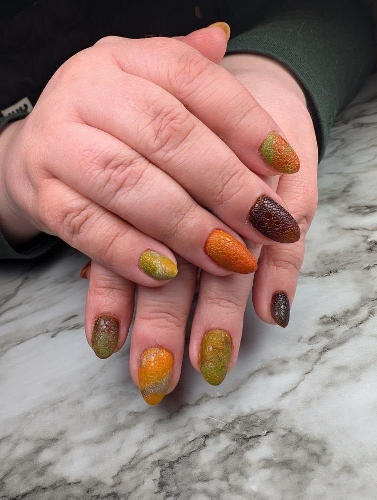 A photo of hands posed to show the fingernails and the nail art on them: the right hand is crossed over the left hand and 8 fingernails are visible (not thumbs). Nails have a scaled pattern on them and are mottled colors of brown, orange, and pale green.