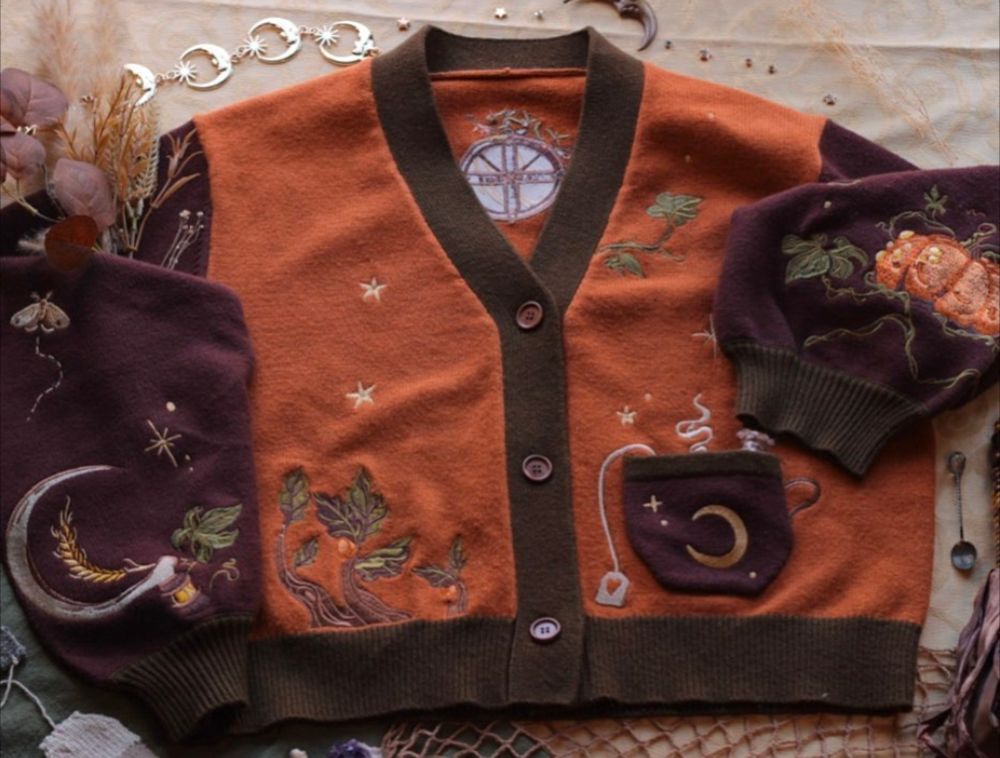 Knock Thrice's photo of the Harvest Witch cardigan laid flat. Cardigan has a pumpkin orange body with olive green trim and cuffs and brown sleeves. The puffed sleeves have embroidery of a pumpkin and vines on one sleeve and wheat, a sickle, vines, and a moth on the other sleeve. There is a small pocket on the right front of the cardigan that is embroidered to look like a steaming mug of tea with a tea bag with a heart on it. The body of the cardigan also features embroidered vines and stars.