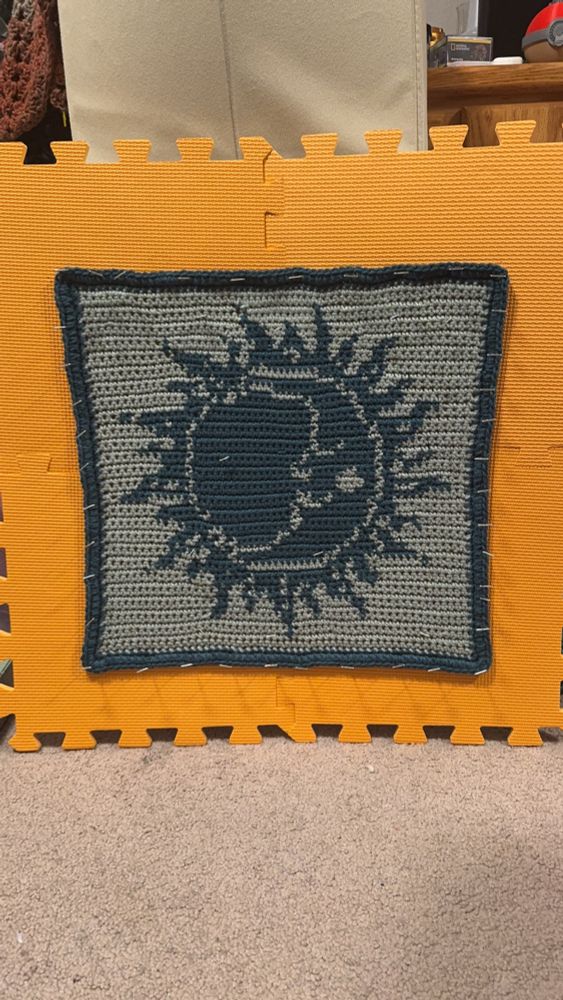 A light sea foam and dark teal crocheted tapestry of a moon inside of a sun with a thick dark teal border.