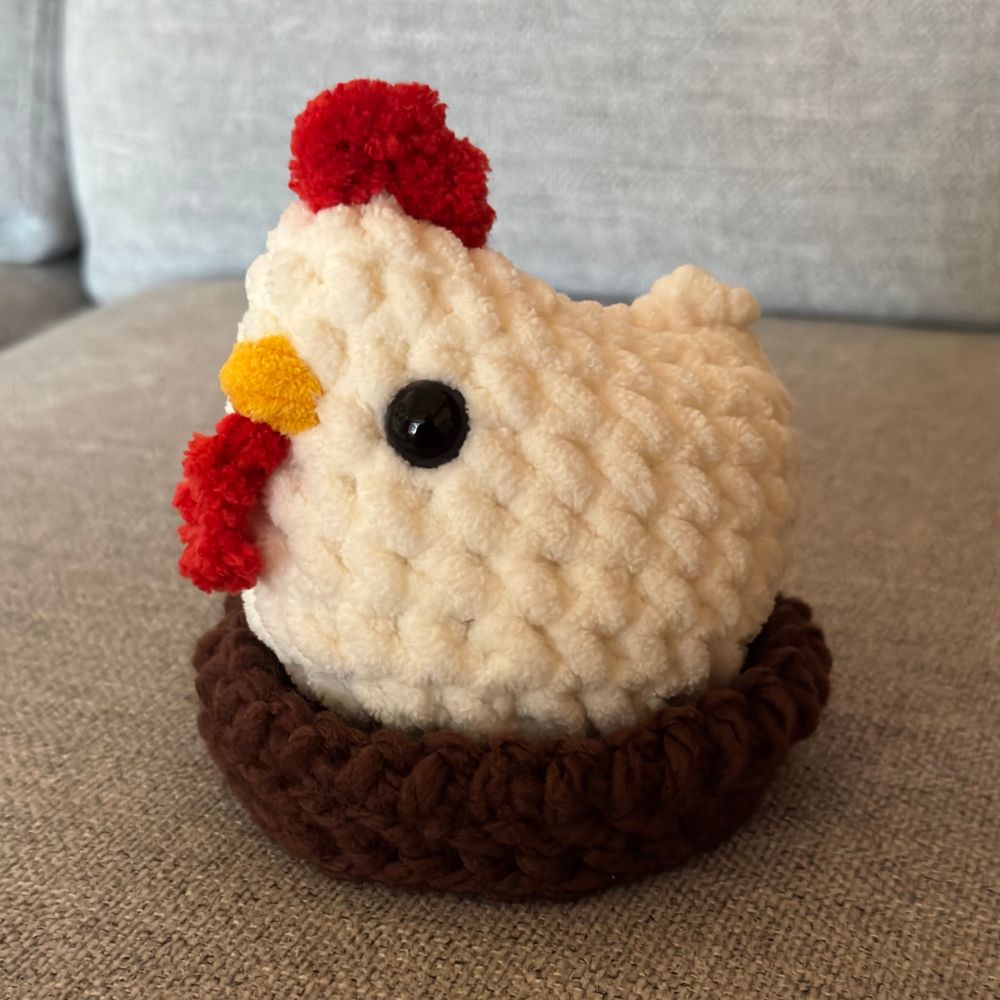 Crocheted chicken in a brown crocheted nest sitting on a light grey couch cushion