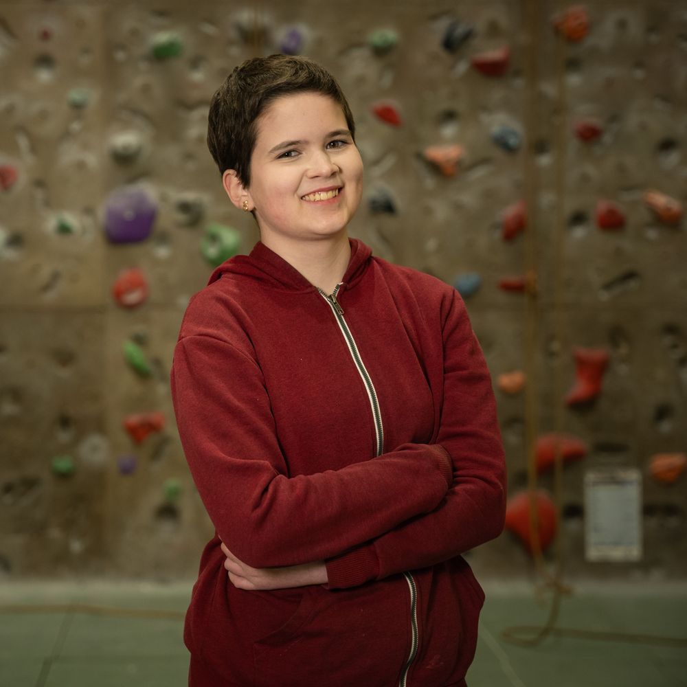 A smiling Rosa, wearing a red zip-up hoody, and standing with her arms crossed in front of a climbing wall.